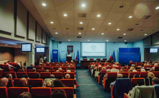 W oddali scena, a tam osoba przemawiająca do uczestników Jubileuszowej Konferencji Okręgu Wielkopolskiego PZN.