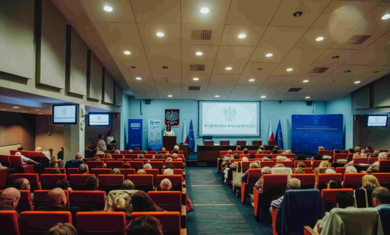 W oddali scena, a tam osoba przemawiająca do uczestników Jubileuszowej Konferencji Okręgu Wielkopolskiego PZN.