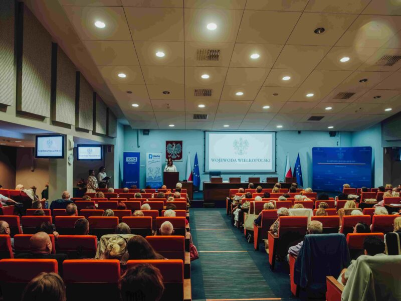 W oddali scena, a tam osoba przemawiająca do uczestników Jubileuszowej Konferencji Okręgu Wielkopolskiego PZN.