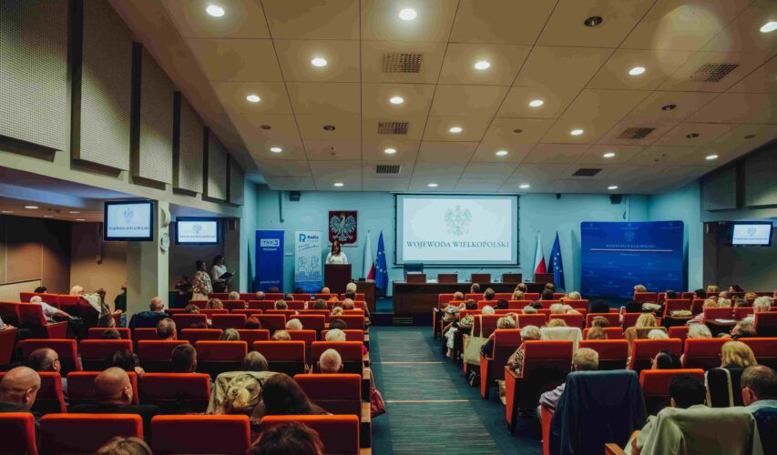 W oddali scena, a tam osoba przemawiająca do uczestników Jubileuszowej Konferencji Okręgu Wielkopolskiego PZN.