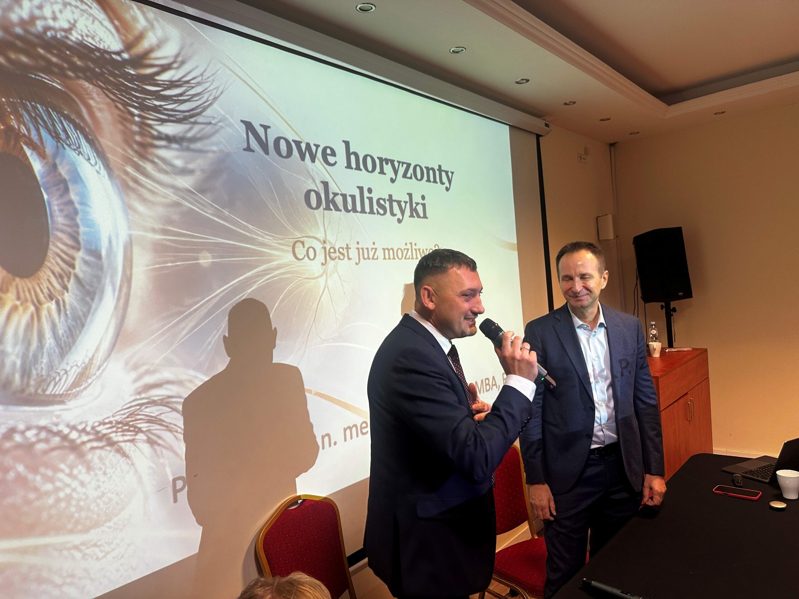Mężczyzna w garniturze trzyma mikrofon. Po jego lewej stronie stoi mężczyzna w garniturze. Za nimi na ścianie ekran ze slajdem z napisem „Nowe horyzonty okulistyki. Co jest już możliwe?”