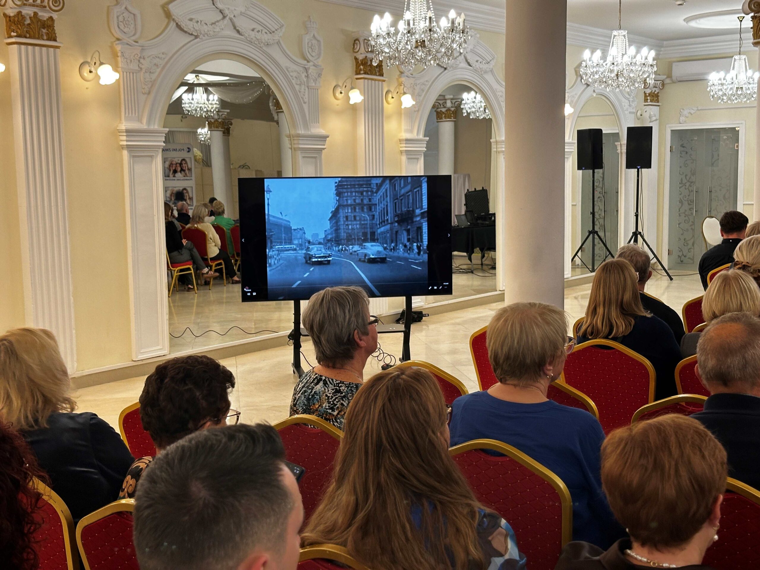 Widoczny fragment sali hotelowej – kilka rzędów z widzami. Widać tyły głów. Z lewej strony ekran z kadrem z czarno-białego filmu. Na nim ulica z samochodami z lat 50., 60. XX w. jadącymi w przeciwnych kierunkach.