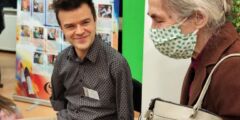 Starsza kobieta rozmawia z uśmiechniętym, młodym mężczyzną, który właśnie szkicuje obraz stopą.