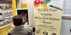 Mężczyzna na wózku inwalidzkim maluje obraz, trzymając w ustach pędzelek. Obok plakat z napisem: Wystawa prac artystów malujących ustami i nogami.
