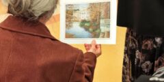 Starsza kobieta trzyma w dłoni papierową reprodukcję obrazu. Zdjęcie zrobione zza jej pleców.