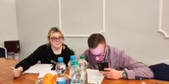 Dwoje młodych ludzi, dziewczyna i chłopak, siedzą przy stole i rozwiązują zadanie związane z planowaniem budżetu. Młody mężczyzna trzyma w dłoni telefon.