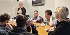 Grupa młodych ludzi siedzi przy stole i słucha prowadzącej.