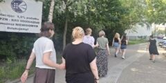Kobiety idą chodnikiem parami. Jedna osoba w każdej parze ma zasłonięte oczy i trzyma swoją przewodniczkę tuż nad łokciem. W tle widać tablicę z logo PZN.