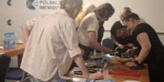 Grupa kobiet stoi wokół stołu i ogląda różne pomoce - maszynę brajlowską, wagę kuchenną, lupy, ramkę do podpisu.